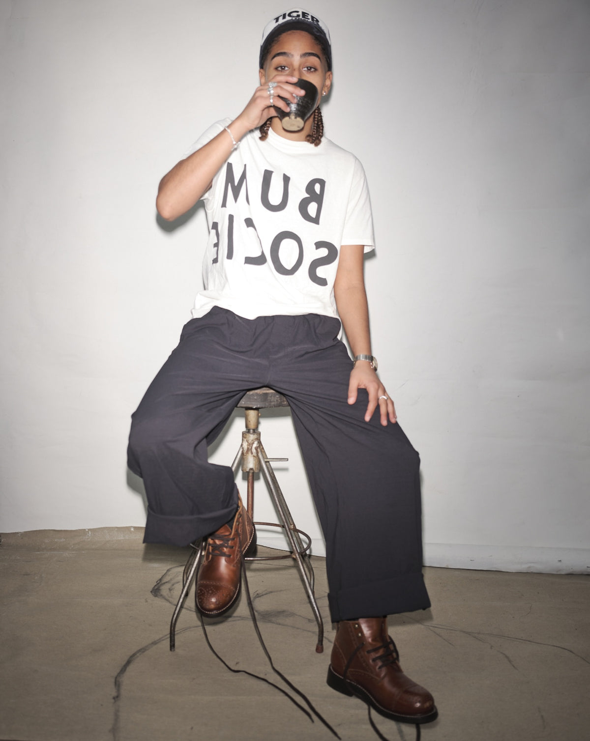Amani Smith sitting on a stool wearing a white t-shirt with Bum Society, dark pants, and brown Bum Society boots.