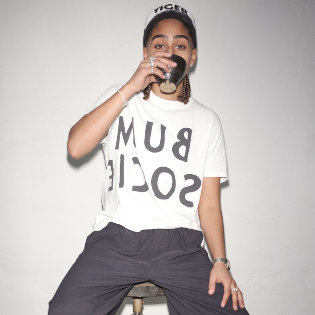 Person wearing a snow reversed Bum Society logo t-shirt and a cap, sitting on a stool against a plain background color:snow