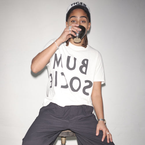 Person wearing a snow reversed Bum Society logo t-shirt and a cap, sitting on a stool against a plain background color:snow