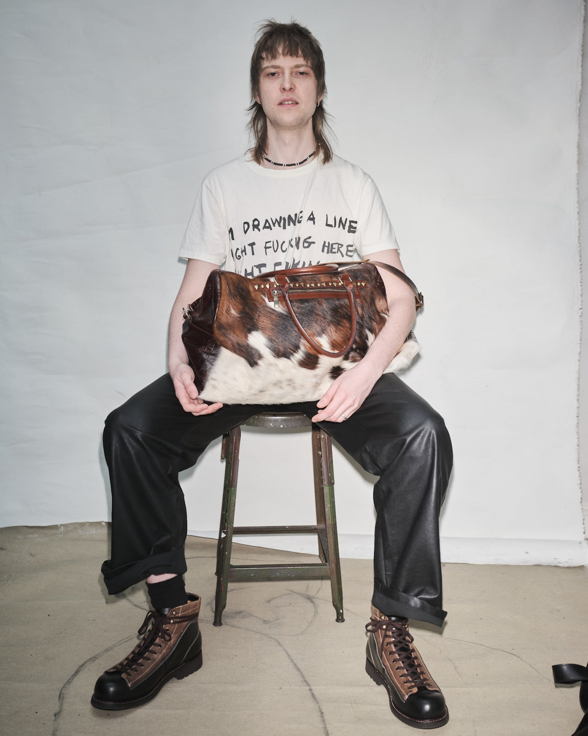 Douglas Diamond sitting on a stool holding a Bum Society pony hair duffle bag against a plain background