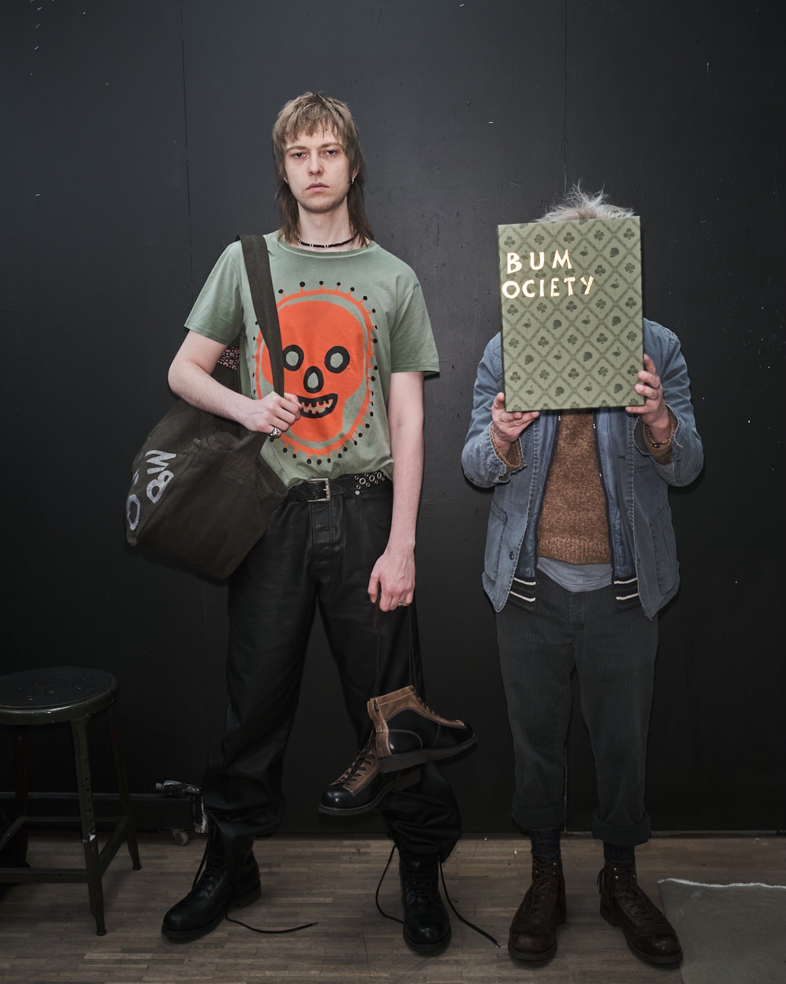 Douglas Diamond in a Waldersten x Bum Society t-shirt  standing against a black wall, holding a Bum Society tote bag and Claes Bondelid holding a Bum Society boot box.