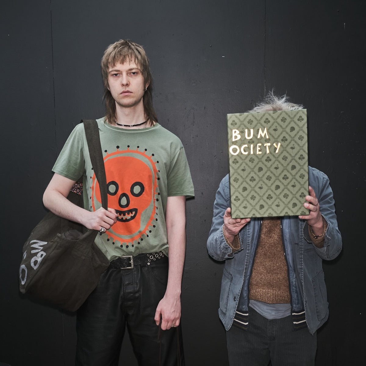 Two people standing against a black background, one holding a bag and the other holding a box labeled 'Bum Society' original artwork by Waldersten for collaboration with Bum Society color:cypress