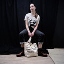 A model sits on a stool in front of a black backdrop, wearing a cream graphic t-shirt and dark jeans, with a cream canvas tote bag featuring black text resting on the floor in front of her.