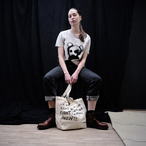 A model sits on a stool in front of a black backdrop, wearing a cream graphic t-shirt and dark jeans, with a cream canvas tote bag featuring black text resting on the floor in front of her.