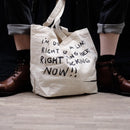 An off-white canvas tote bag with black handwritten text sits on the floor between a person's legs, who are wearing rolled-up dark jeans and brown leather boots.