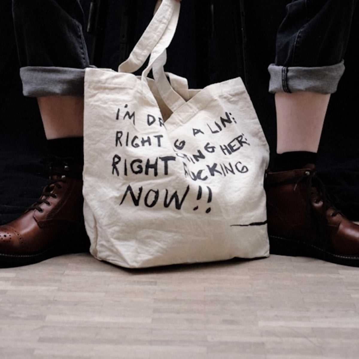 An off-white canvas tote bag with black handwritten text sits on the floor between a person's legs, who are wearing rolled-up dark jeans and brown leather boots.