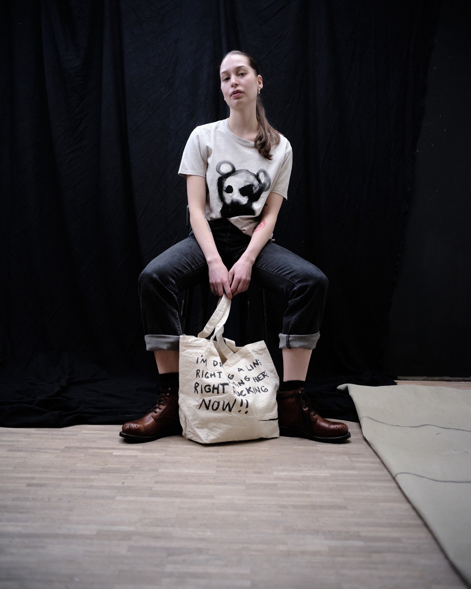 Elsa Myrgard sitting on a stool holding a Waldersten x Bum Society tote bag with text against a black curtain background