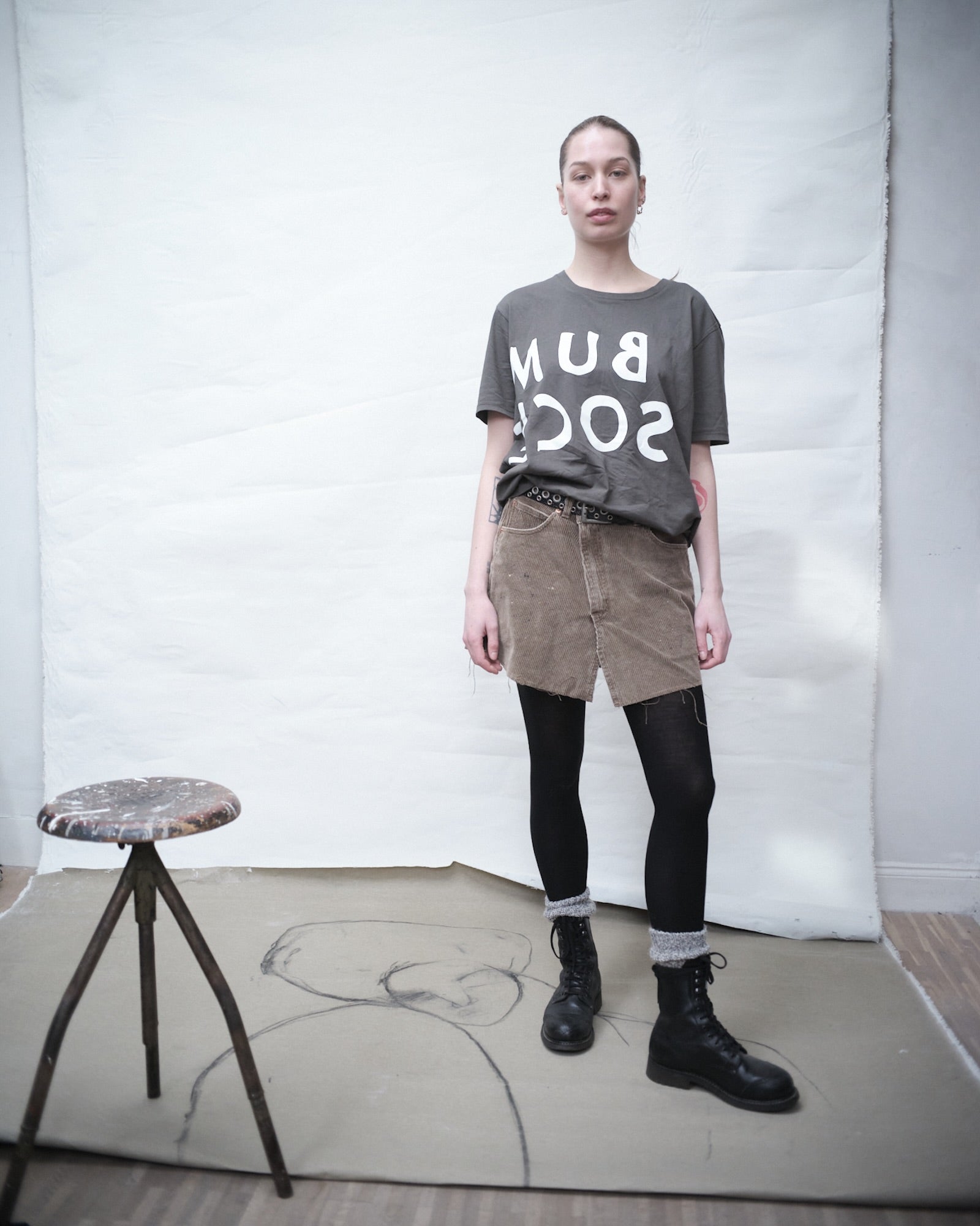 Elsa Myrgard wearing an ink Bum Society t-shirt with logo, beige skirt, black leggings, and black Bum Society boots in a minimalistic setting.
