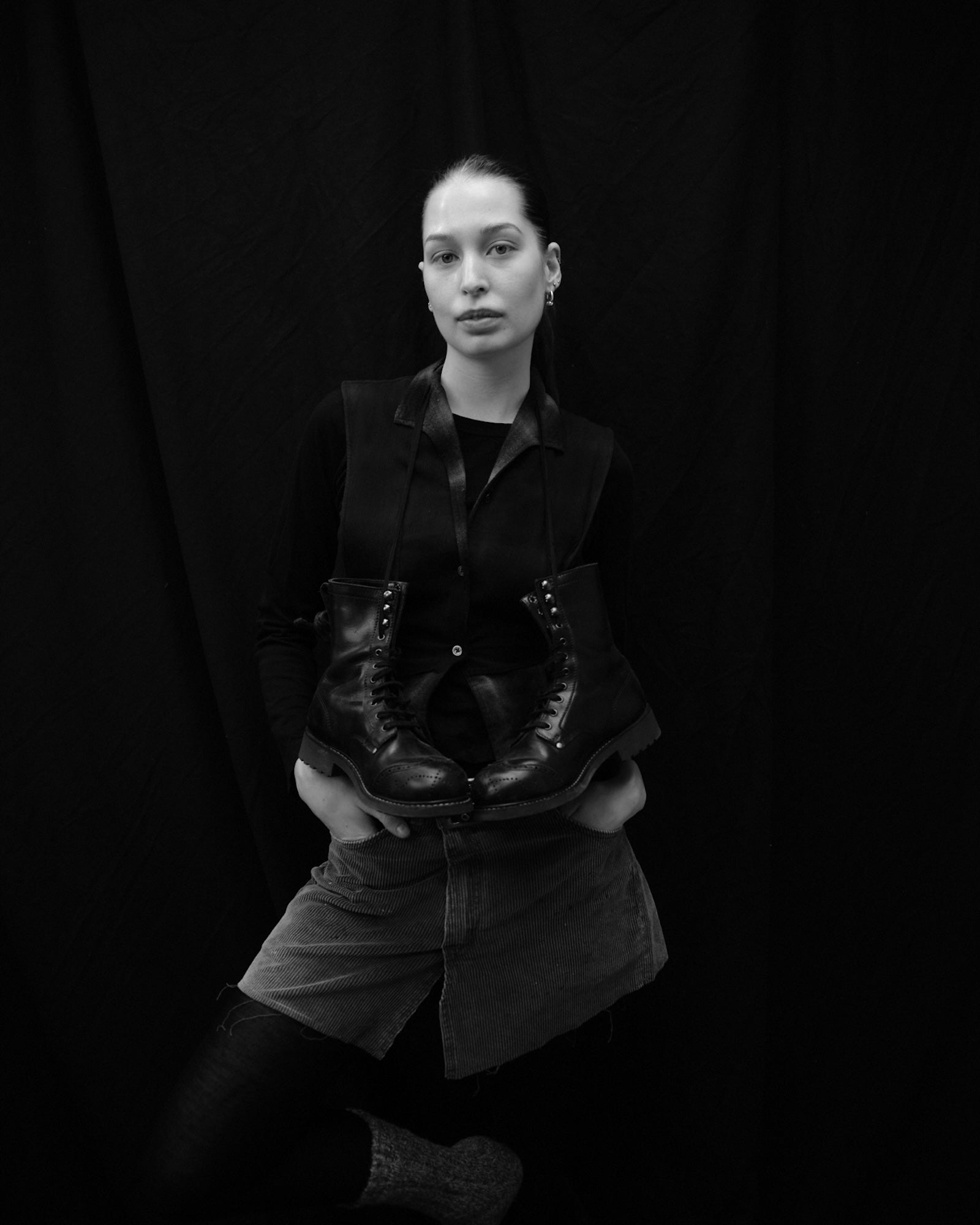 Black and white portrait of Elsa Myrgard wearing a dark jacket and shorts against a black background holding a pair of Bum Society boots