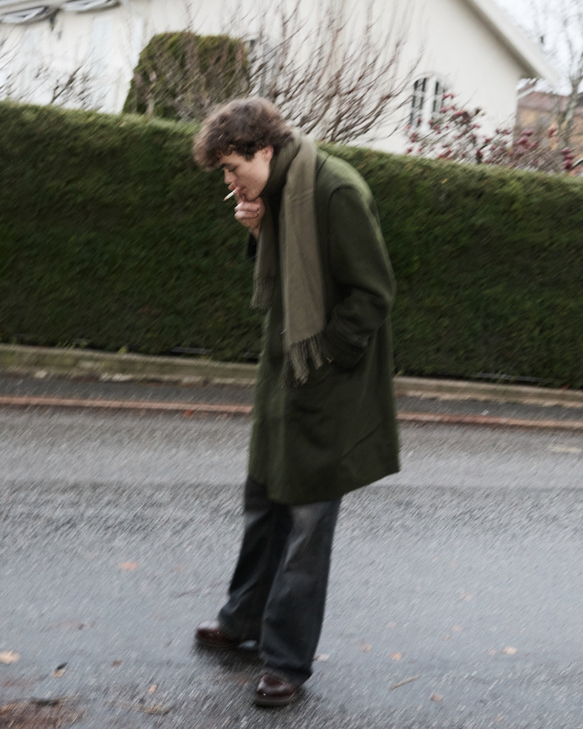 Person wearing a green coat and scarf standing on a street with a hedge and building in the background.