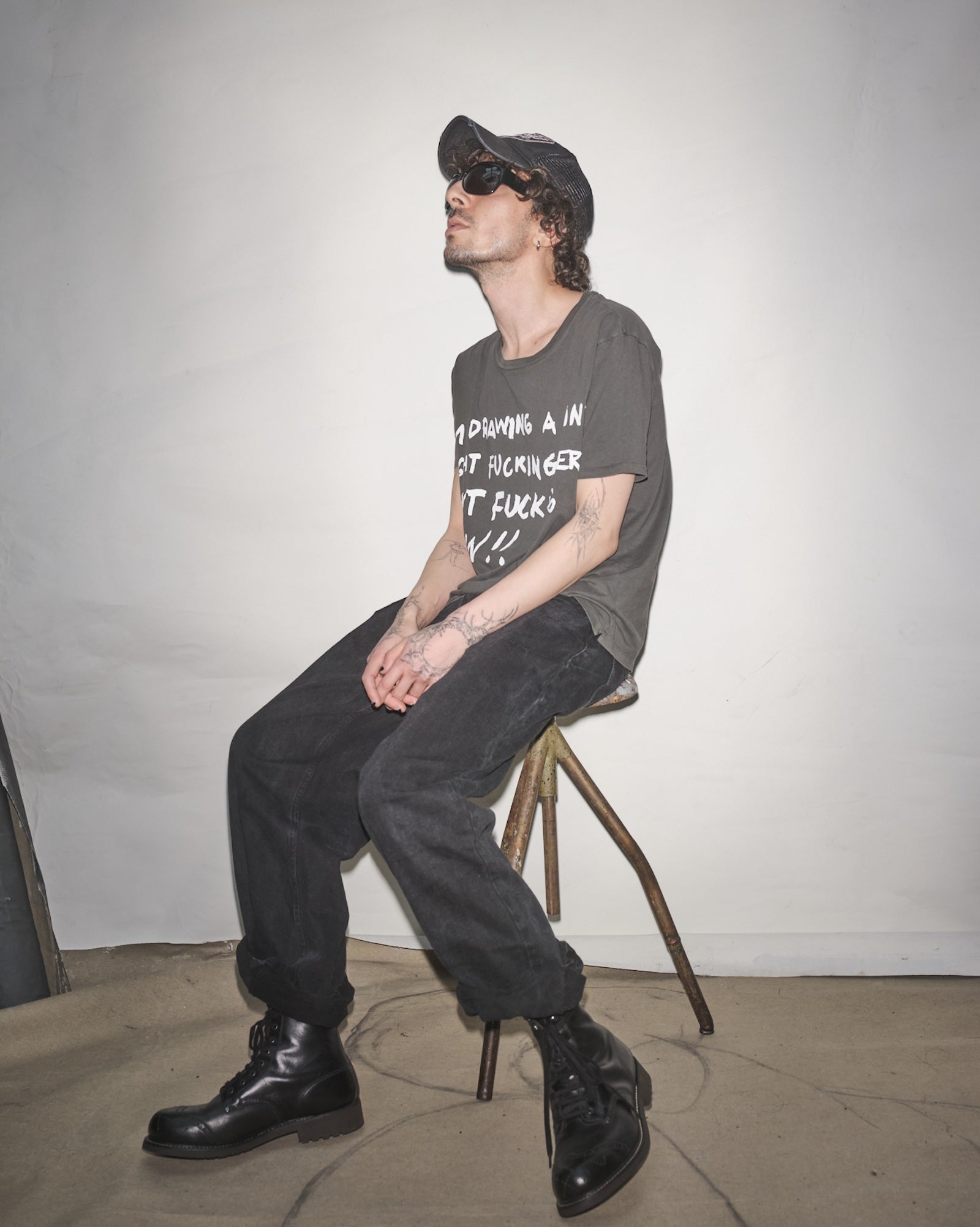 Josef Forselius sitting on a stool wearing a Bum Society t-shirt, black pants, and black Bum Society boots against a plain background.