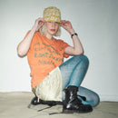 Person wearing an orange Waldersten x Bum Society t-shirt, blue patterned leggings, and black Bum Society boots sitting on a white floor color:bs orange