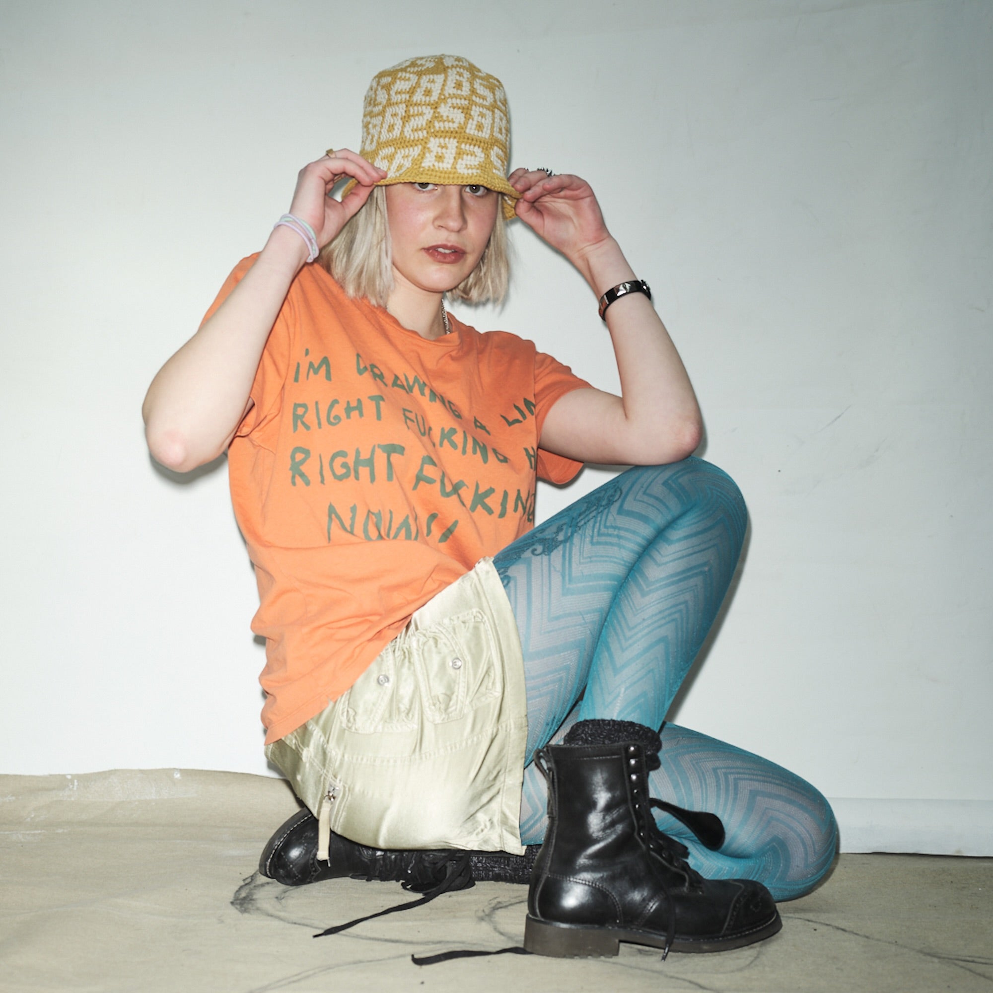 Person wearing an orange Waldersten x Bum Society t-shirt, blue patterned leggings, and black Bum Society boots sitting on a white floor color:bs orange
