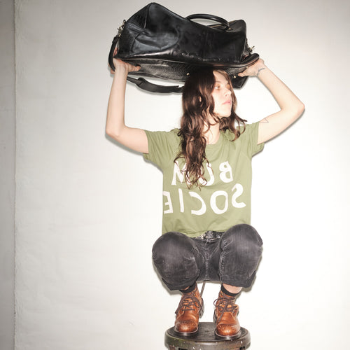 Person wearing a green t-shirt with text, holding a Bum Society leather duffle bag over their head against a plain background color:shiny black