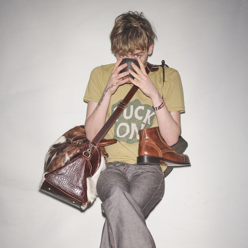 Person sitting with a Bum Society pony hair duffle bag and Bum Society boots, wearing an Ongoing t-shirt in color Earth color:earth