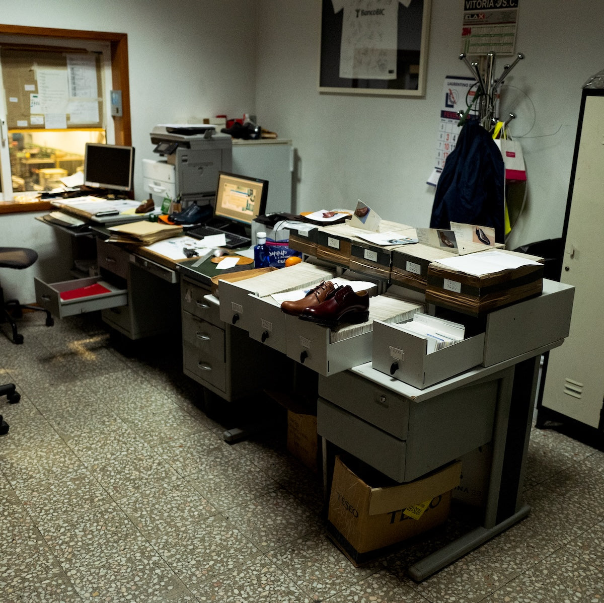 Office desk with computer, shoes, and supplies in a room with a window to the factory