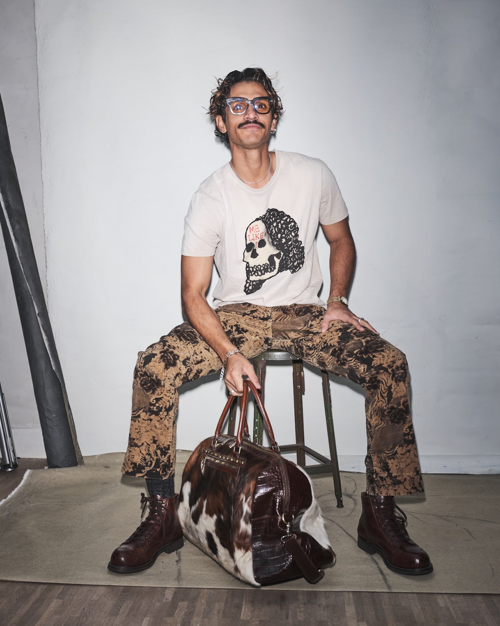 Wazeer Hamdulay sitting on a stool with a Bum Society pony hair duffle bag, wearing a Waldersten x Bum Society t-shirt and camouflage pants.