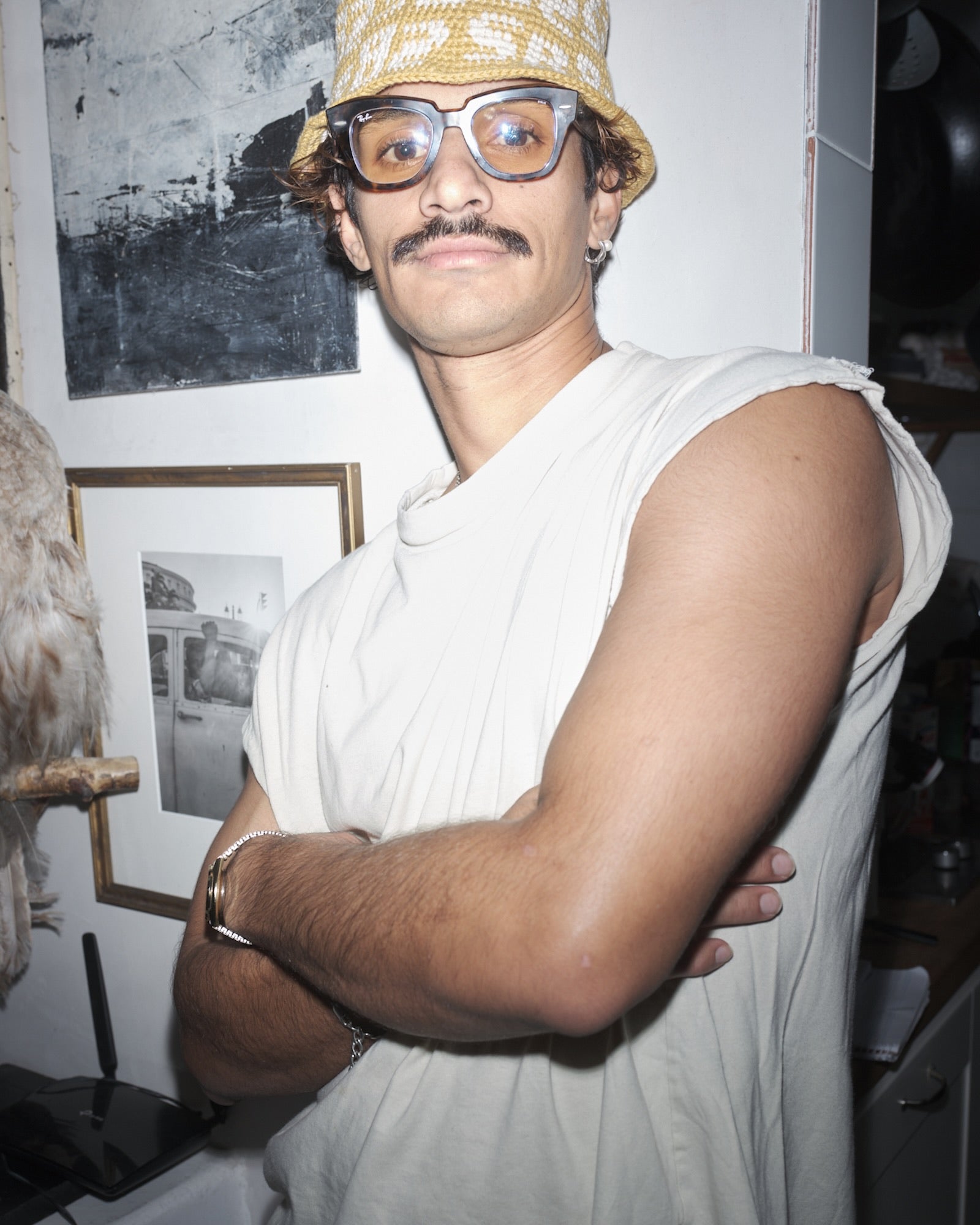 Wazeer Hamdulay wearing a white sleeveless shirt and Bum Society crochet bucket hat  indoors with framed pictures on the wall.